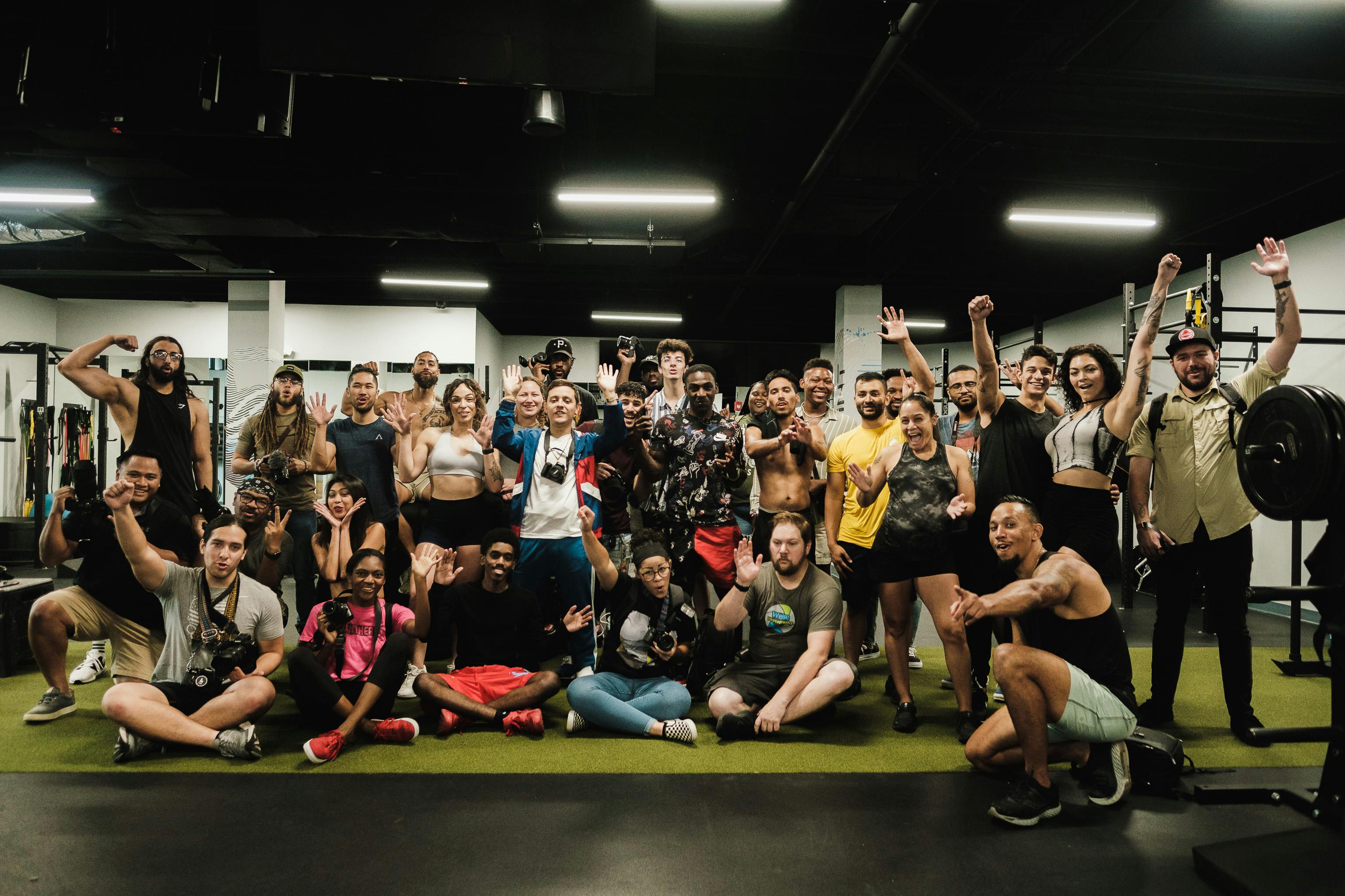 A large group of diverse people smiles and poses energetically in a modern gym setting.